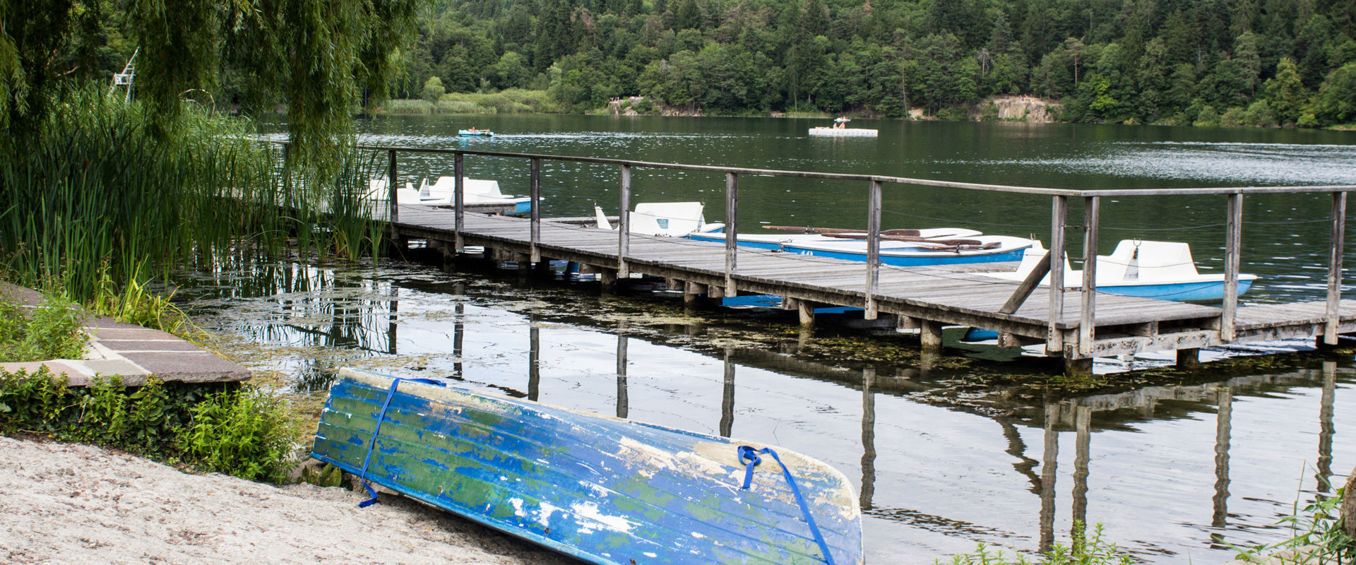 A blue boat lies on the sandy shore of Lake Montiggl. The footbridge in the background with light blue pedal boats leads into the lake.