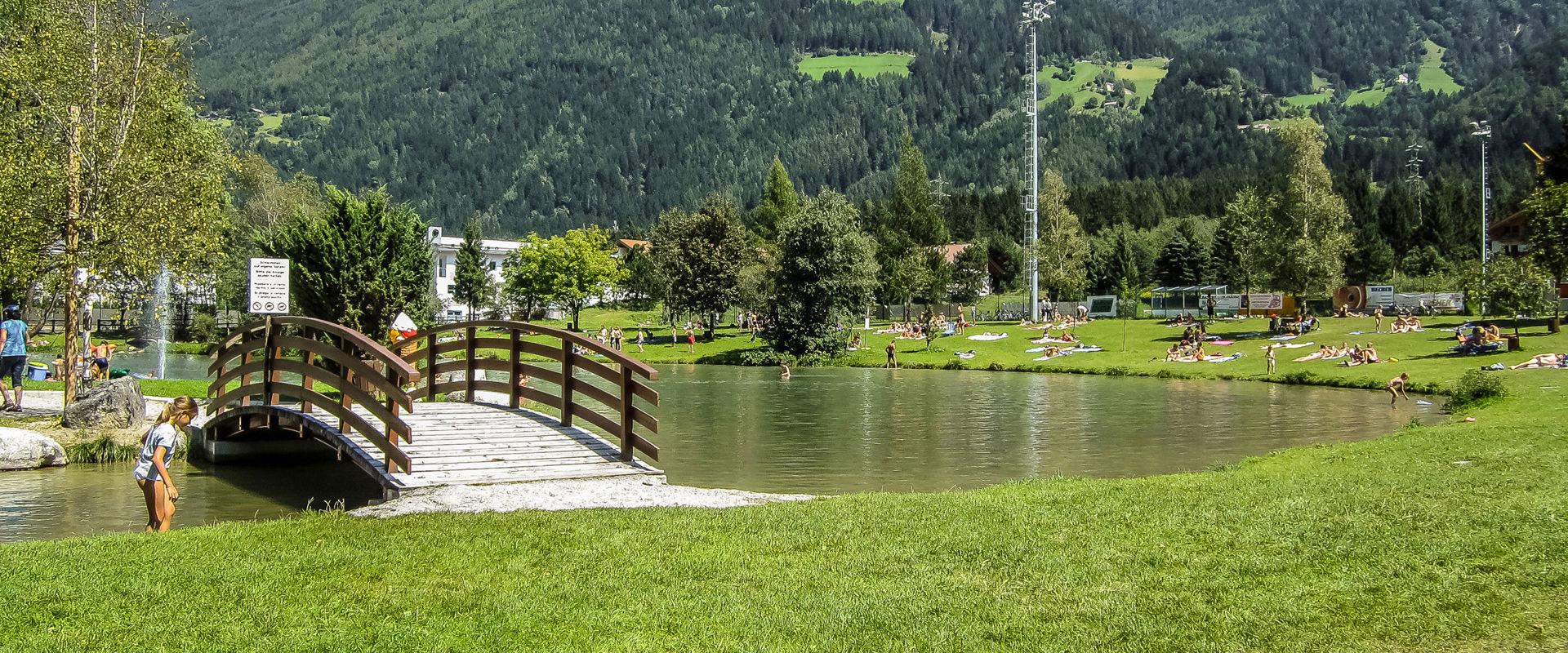 Romantic bathing lake with a small bridge in the middle of greenery and surrounded by mountains.
