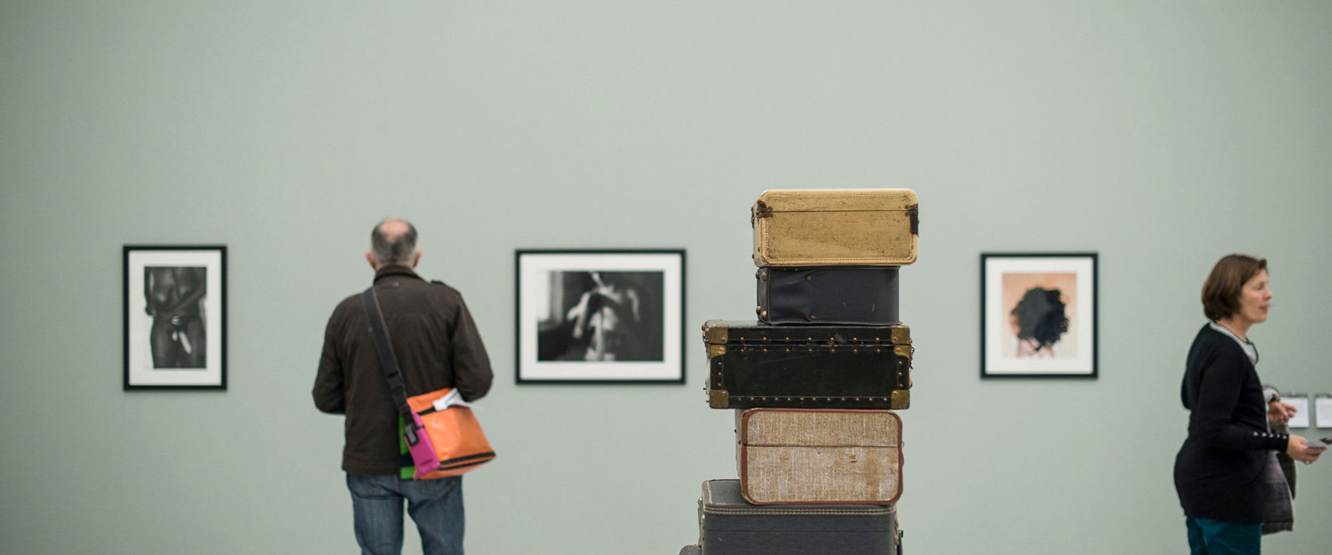 Museion Several old suitcases lie on top of each other in the middle of the room. Pictures hang on the walls and are being examined by 2 visitors.