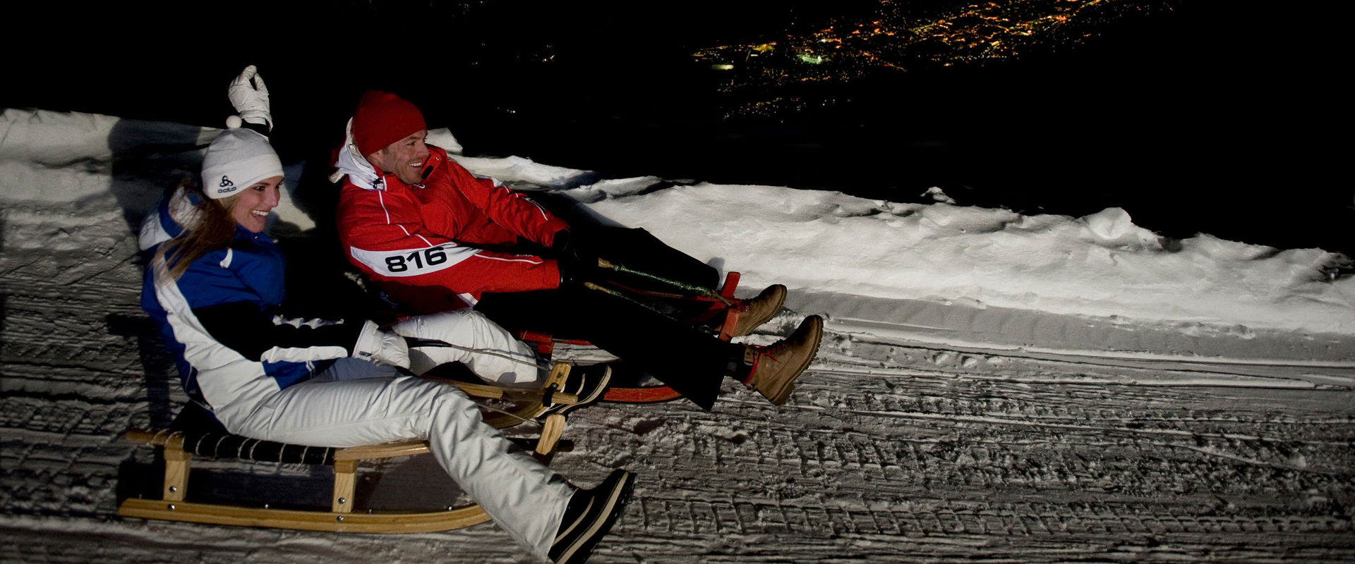 Night tobogganing Two tobogganists ride down the slope at night. In the background, you can see village lights.
