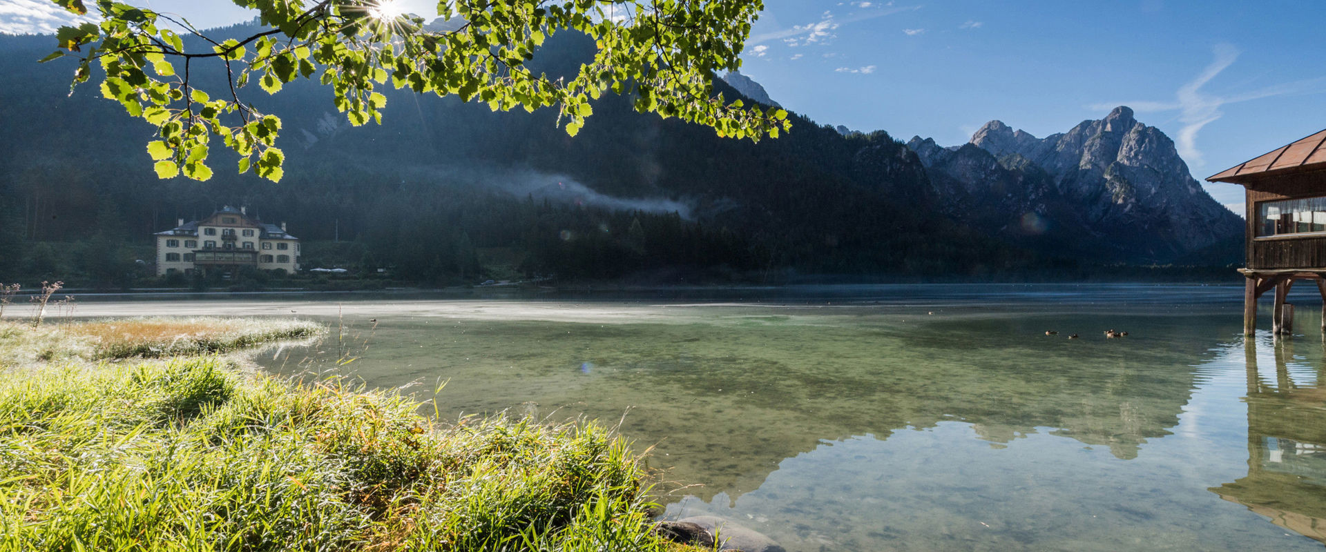 Shore of a clear lake in the mountains, building in the background.