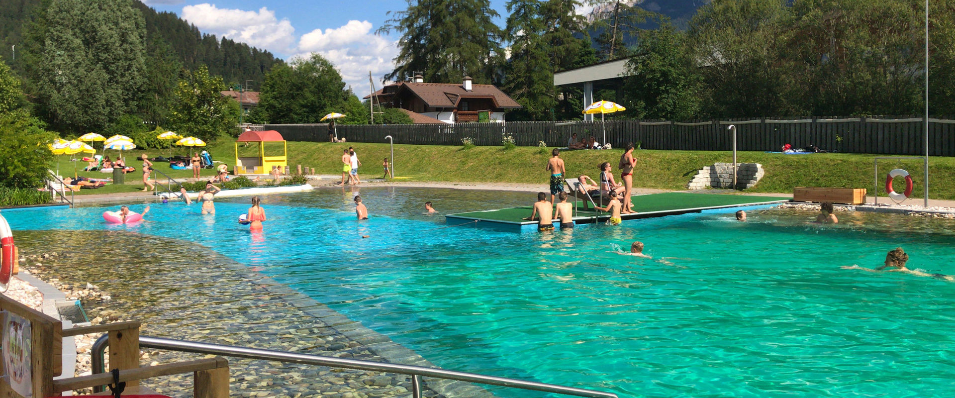 Bathing lake with kids swimming area, bridge, showers and sunbathing lawn. In the background you can see houses and trees.