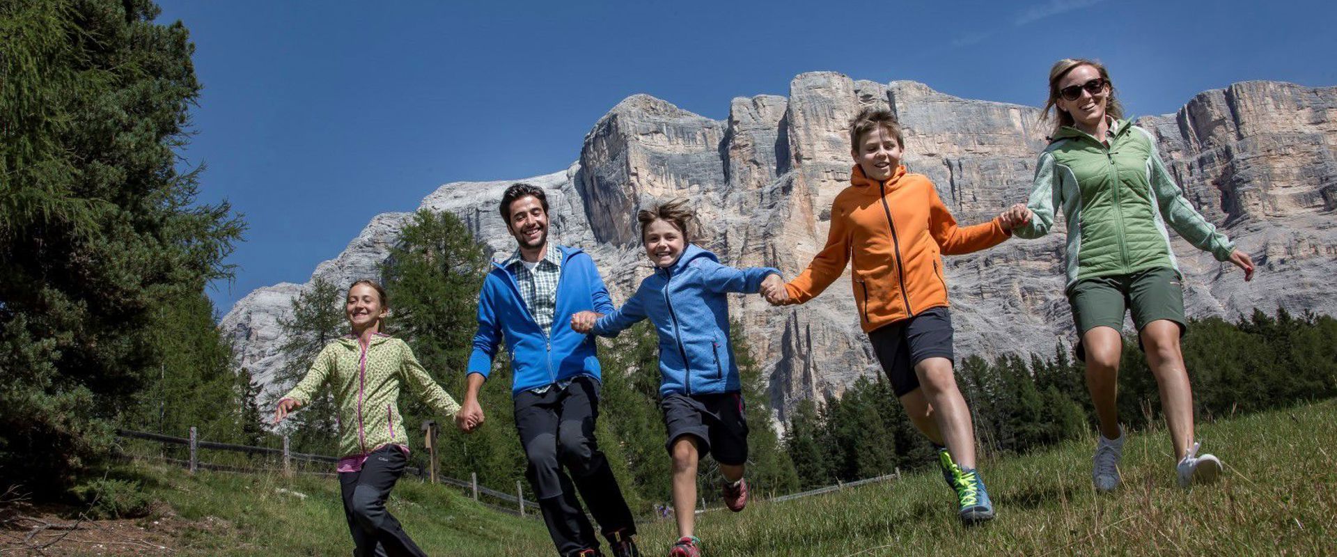 Family holding hands and running on a meadow. Steep rock faces can be seen in the background.
