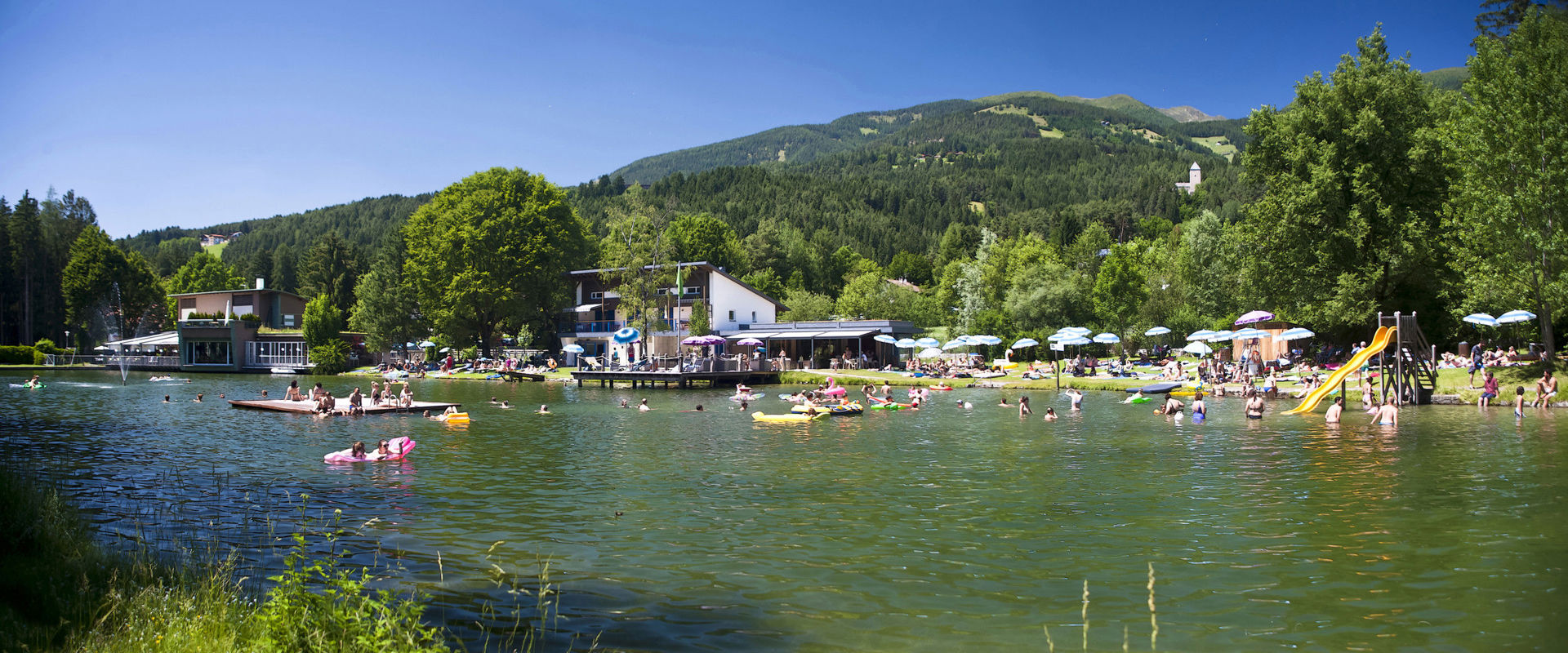 Bathing lake with restaurant, jetty, raft, slide and blue and white parasols.