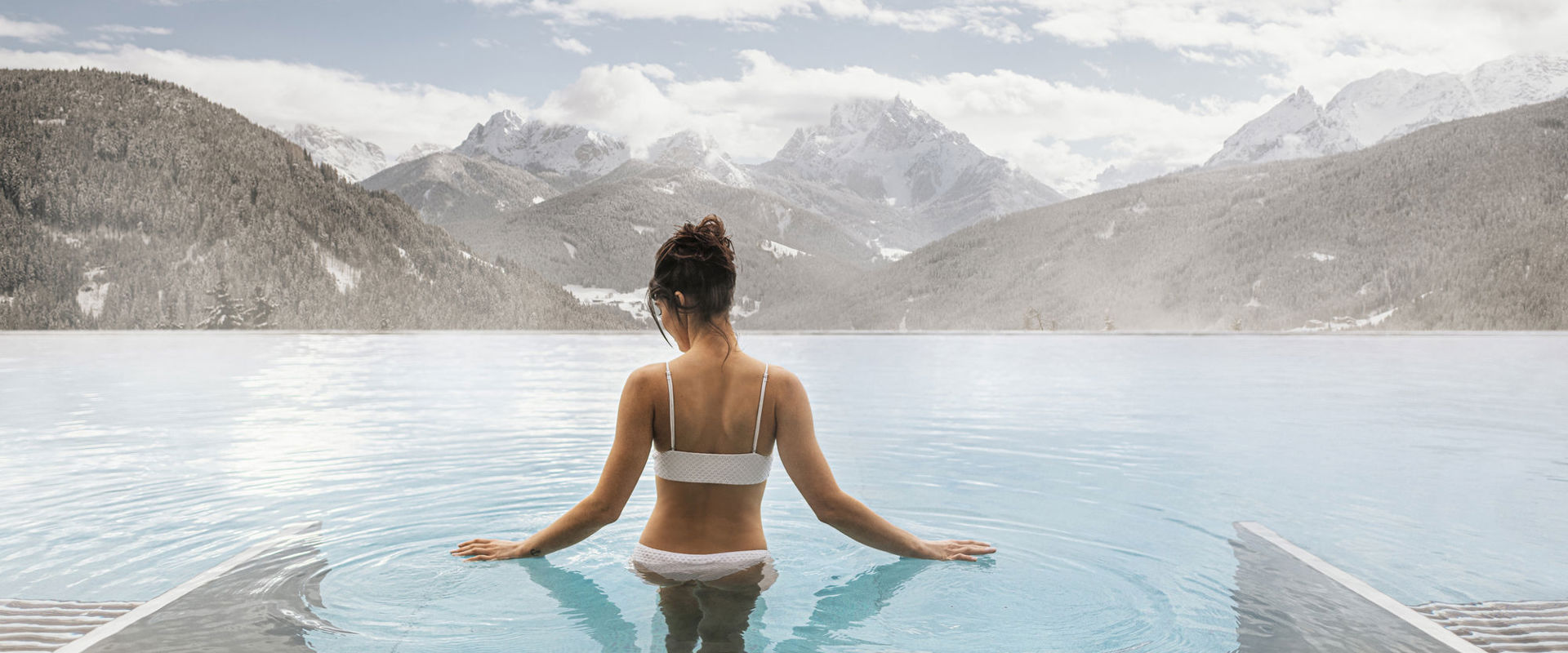 Wellness outdoor pool  Woman wearing a white bikini and with dark hair tied up, descends the steps into the steaming outdoor pool. In the background, you can see the snow-covered mountains.